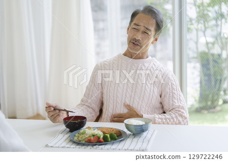 A man in his 60s with a painful expression on his face, holding his stomach at the dinner table A man in his 60s with a painful expression on his face, holding his stomach at the dinner table 129722246