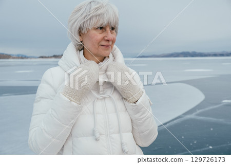 older woman enjoys winter moment on frozen lake, dressed warmly in white attire and mittens. closeup 129726173