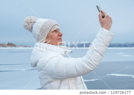 woman takes winter selfie by icy lake with snow covered landscape in background. closeup. 129726177