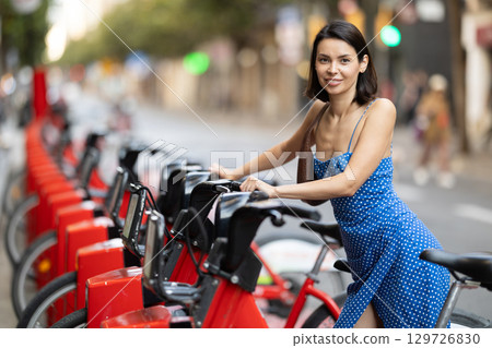 Brunette in blue dress rents electric bike on street of european city Brunette in blue dress rents electric bike on street of european city 129726830