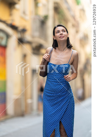 Cheerful brunette girl in blue dress walking along the street among architecture with bag Cheerful brunette girl in blue dress walking along the street among architecture with bag 129726919