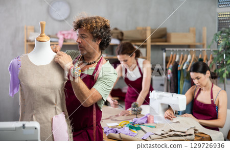 Middle-aged male dressmaker working with mannequin while others sewing and drawing with curves Middle-aged male dressmaker working with mannequin while others sewing and drawing with curves 129726936
