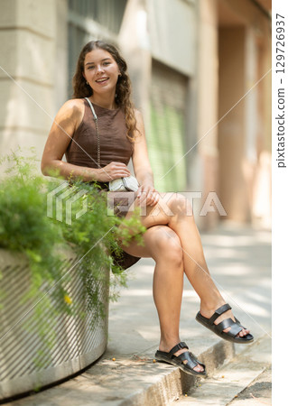 Beautiful young lady wearing dress walking shady street and sitting concrete flower bed with plants, taking break from heat of day Beautiful young lady wearing dress walking shady street and sitting concrete flower bed with plants, taking break from heat of day 129726937