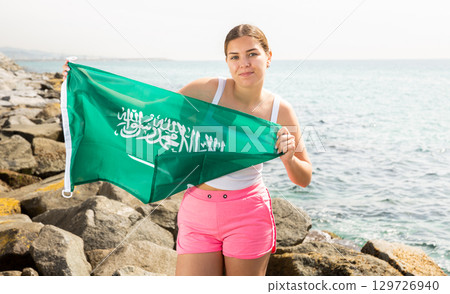 Young woman on coast posing with flag of Saudi Arabia Young woman on coast posing with flag of Saudi Arabia 129726940