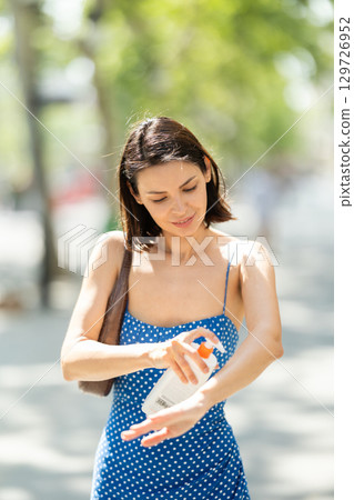 Young brunette girl applying sunscreen to her skin while walking in city Young brunette girl applying sunscreen to her skin while walking in city 129726952