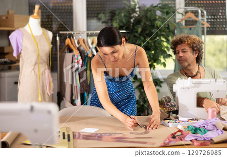 Young female dressmaker drawing with curves while man sewing Young female dressmaker drawing with curves while man sewing 129726985