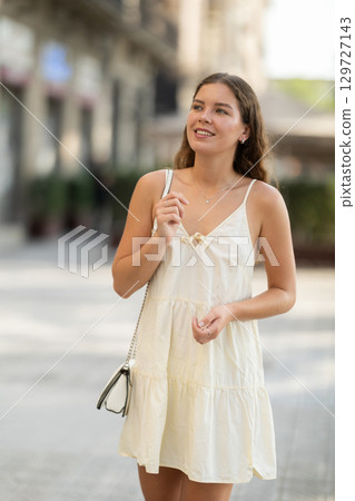 Cheerful girl in white dress walking along the street among architecture with bag Cheerful girl in white dress walking along the street among architecture with bag 129727143