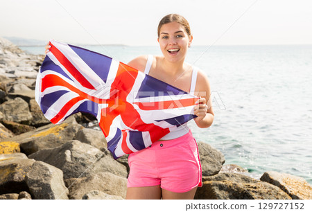 Attractive smiling woman holding Great Britain flag on the seashore on sunny day 129727152