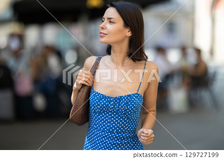 Cheerful brunette girl in blue dress walking along the street among architecture with bag 129727199