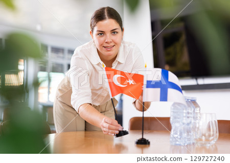 Positive young woman putting flag of Turkey on the table with flag of Finland in conference room 129727240