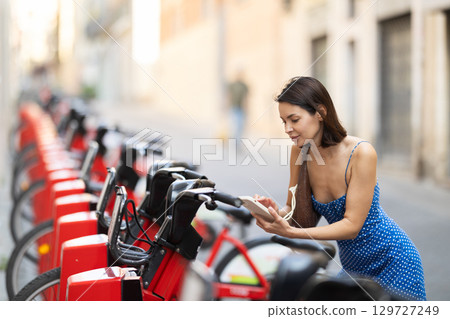 Brunette in blue dress rents electric bike - pays with mobile phone at street parking Brunette in blue dress rents electric bike - pays with mobile phone at street parking 129727249