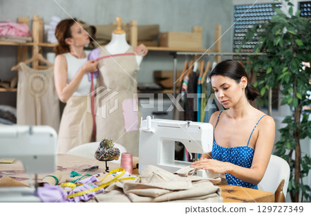 Seamstress working on modern sewing machine in sewing workshop. Dressmaker tries on a dress on mannequin 129727349
