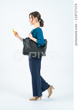 Full body photo of a white woman walking while looking at her smartphone 129727374