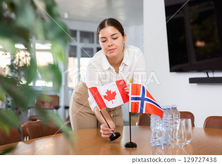Positive young woman putting flag of Canada on the table with flag of Norway in conference room Positive young woman putting flag of Canada on the table with flag of Norway in conference room 129727438