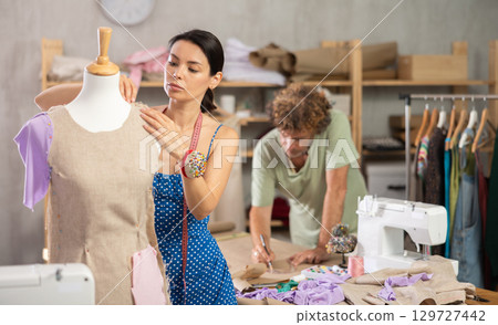 Young female dressmaker working with mannequin while assistant drawing fashion sketch 129727442