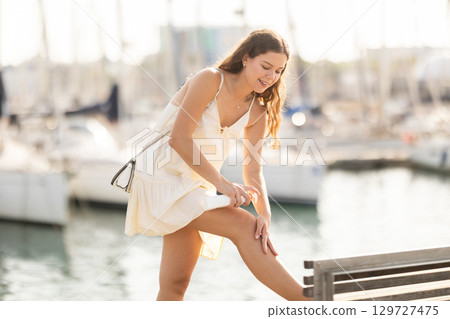 Young woman applying sunscreen on bench near port 129727475
