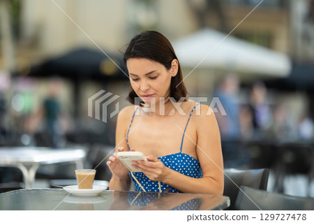 Brunette texting on smartphone screen during breakfast on cafe terrace. Drinking coffee 129727478
