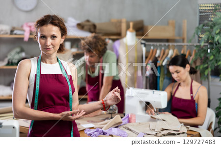 Middle-aged female dressmaker inviting clients while others sew and draw Middle-aged female dressmaker inviting clients while others sew and draw 129727483