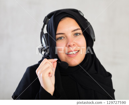 smiling Muslim woman in black hijab and headphones with microphone on gray background. 129727590