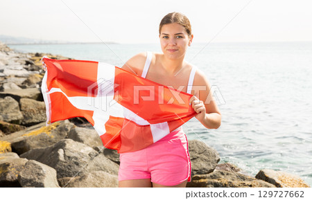 Pretty young girl holding the Danish flag in her hands on the seashore on sunny day Pretty young girl holding the Danish flag in her hands on the seashore on sunny day 129727662