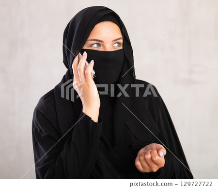 Young woman dressed in traditional islamic clothes with her face covered having phone conversation Young woman dressed in traditional islamic clothes with her face covered having phone conversation 129727722