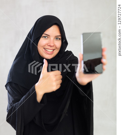 smiling Muslim woman in traditional black hijab shows blank smartphone screen close-up 129727724