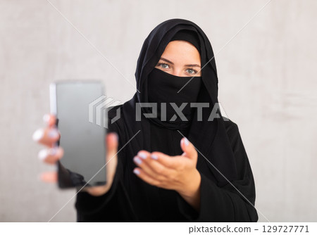 young Muslim girl with her face covered by burka shows empty dark smartphone screen. 129727771