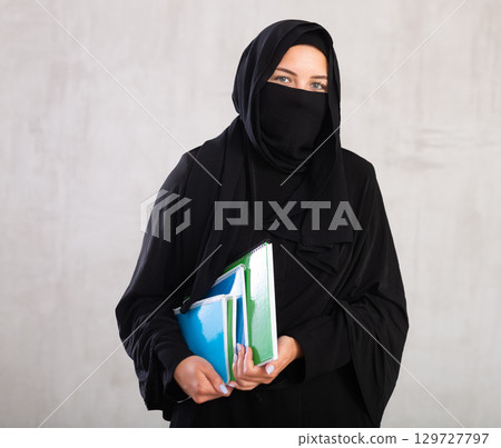 Young student woman dressed in traditional islamic clothes with her face covered holding notebooks Young student woman dressed in traditional islamic clothes with her face covered holding notebooks 129727797