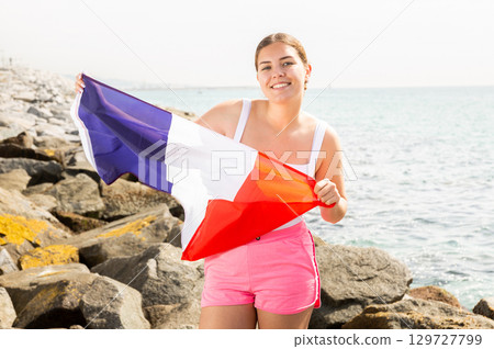 Young happy woman holding flag of French with a happy face and smiling on seashore on sunny day 129727799