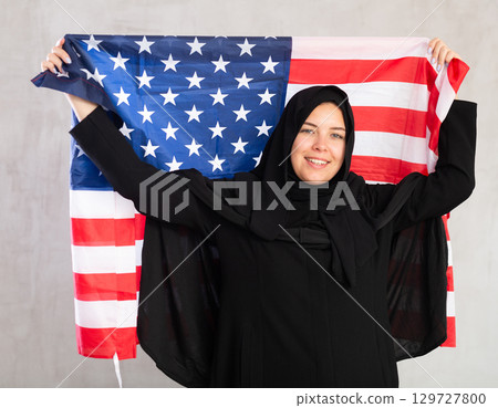 Balanced smiling Muslim woman in traditional black hijab holds flag of United States of America 129727800