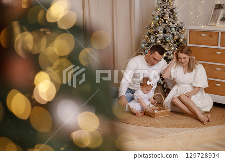 Young family with baby playing near Christmas tree in cozy room with white wall and wooden floor 129728934