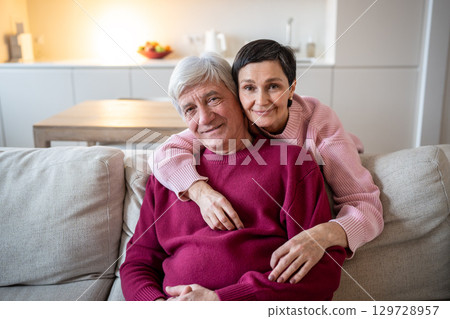 Happy elderly couple hugging on sofa at home smiling and looking at camera. Love on retirement Happy elderly couple hugging on sofa at home smiling and looking at camera. Love on retirement 129728957