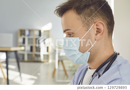 Close up portrait of pensive man doctor or nurse in blue uniform and face mask standing in office. 129729893
