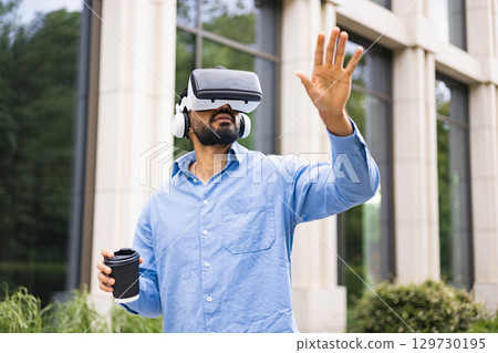 A man wearing a VR headset and headphones, reaching out while holding a coffee cup outdoors near a building. 129730195