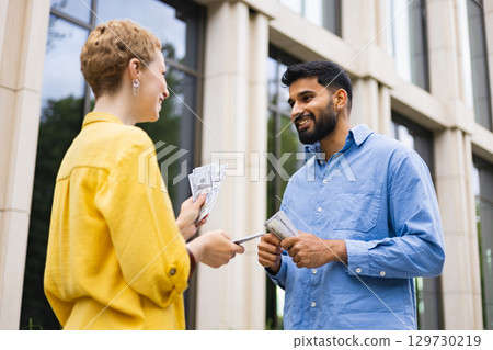 A woman hands money to a man outdoors, likely a financial transaction, against a building. A woman hands money to a man outdoors, likely a financial transaction, against a building. 129730219