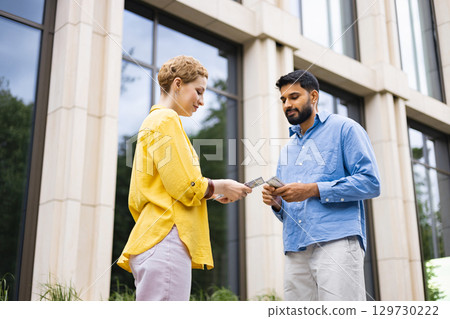 A woman in a yellow shirt hands money to a man in a blue shirt, both outdoors in front of a building. A woman in a yellow shirt hands money to a man in a blue shirt, both outdoors in front of a building. 129730222