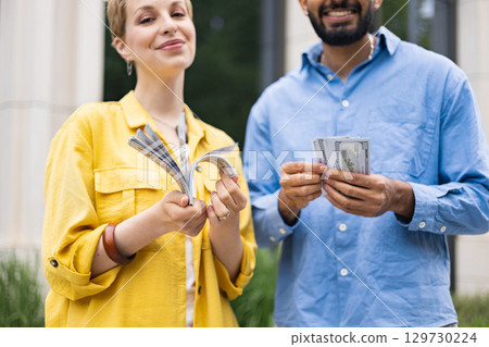 A diverse couple joyfully holds stacks of American hundred dollar bills, symbolizing financial success and wealth. A diverse couple joyfully holds stacks of American hundred dollar bills, symbolizing financial success and wealth. 129730224
