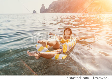 Woman, Inflatable, Sea - Woman floats on inflatable raft in calm ocean water with rocky cliffs in the distance. 129732839