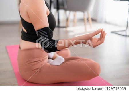 Woman sitting cross legged on yoga mat in lotus pose wearing sportswear focusing on breathing in modern apartment Woman sitting cross legged on yoga mat in lotus pose wearing sportswear focusing on breathing in modern apartment 129732939