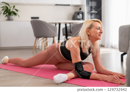 Focused blonde middle-aged woman performing single-leg stretch on floor mat in modern living room for flexibility and wellness. 129732942