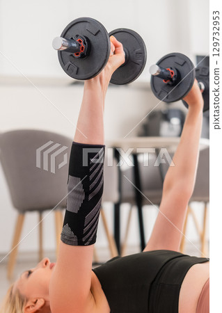 Blonde woman in middle age exercising chest press with dumbbells on yoga mat, training for strength and health in home interior 129732953