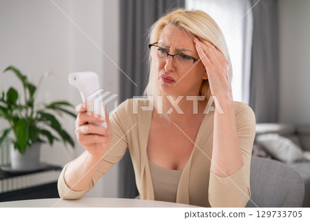 Blonde middle aged woman in beige outfit looking worried at thermometer, experiencing high temperature and discomfort in modern living room Blonde middle aged woman in beige outfit looking worried at thermometer, experiencing high temperature and discomfort in modern living room 129733705