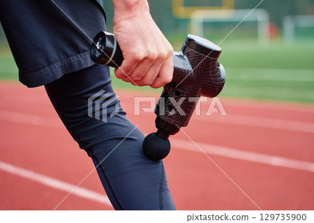 Athlete massaging leg with massage gun on running track 129735900
