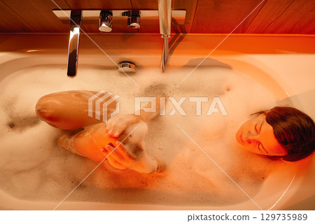 Woman taking bubble bath in cozy bathroom with romantic lighting 129735989