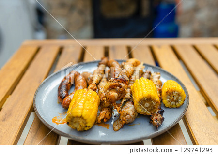 Grilled sausages, skewers, and corn on a plate near a gas barbecue with vegetables and flatbreads cooking. Outdoor summer cooking scene with delicious grilled food. Grilled sausages, skewers, and corn on a plate near a gas barbecue with vegetables and flatbreads cooking. Outdoor summer cooking scene with delicious grilled food. 129741293