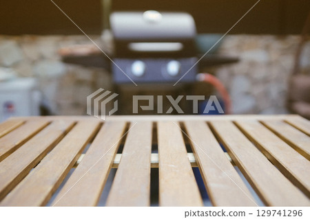 Close-up of a wooden outdoor table with a blurred gas grill in the background. Backyard BBQ setup ready for summer cooking and social gatherings. Close-up of a wooden outdoor table with a blurred gas grill in the background. Backyard BBQ setup ready for summer cooking and social gatherings. 129741296