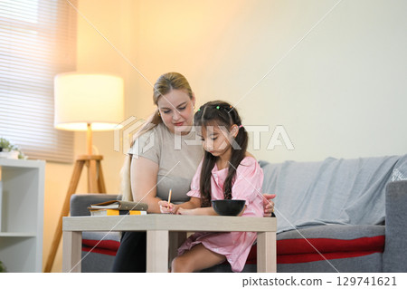 Caring mother helping her daughter with homework at home, sitting together in the living room Caring mother helping her daughter with homework at home, sitting together in the living room 129741621