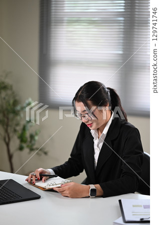 Female worker in black suit writing notes in a planner while working at office desk 129741746