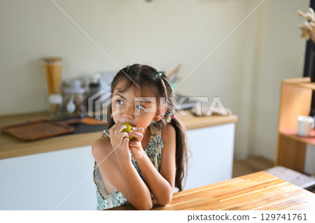 Adorable girl with pigtails eating fresh fruit at the wooden table during breakfast Adorable girl with pigtails eating fresh fruit at the wooden table during breakfast 129741761