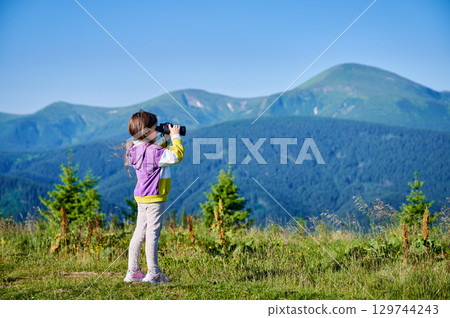 Young girl stands in grassy meadow, looking through binoculars, wearing yellow and purple sweatshirt. Behind her, rolling green hills and mountains stretch into distance under clear blue sky. 129744243